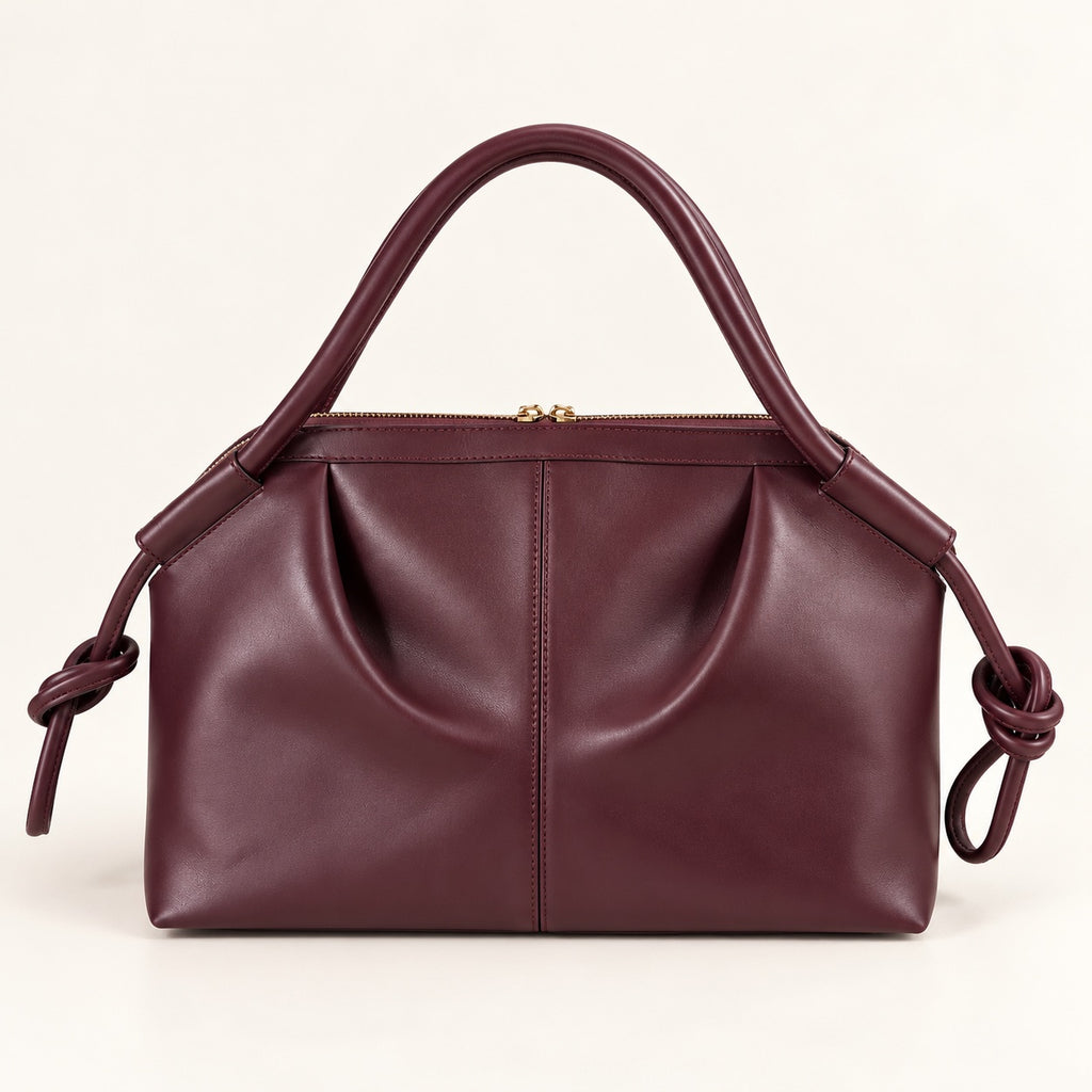 A burgundy leather handbag with a top handle, a top zipper closure, and knotted leather strap details on the sides, shown against a plain off-white background.