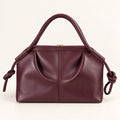 A burgundy leather handbag with a top handle, a top zipper closure, and knotted leather strap details on the sides, shown against a plain off-white background.