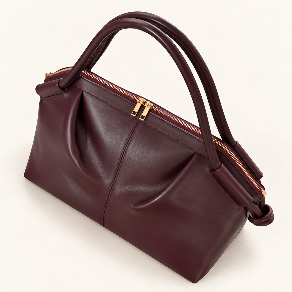 A dark burgundy leather handbag with dual top handles, gold-tone hardware, and a zippered top closure, photographed on a clean off-white background.