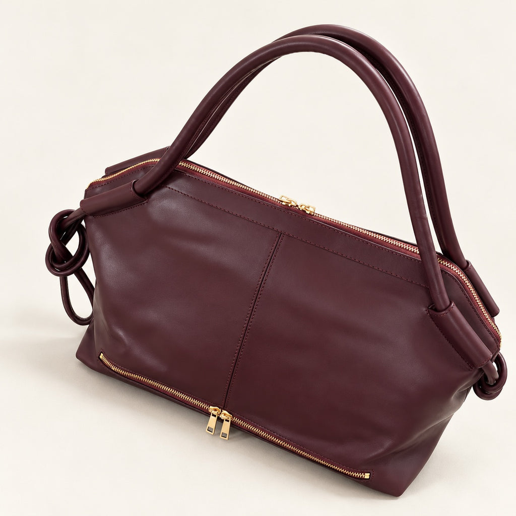 A structured burgundy leather handbag with dual top handles, gold-tone zipper details, and a matching burgundy shoulder strap, photographed on a clean off-white background.