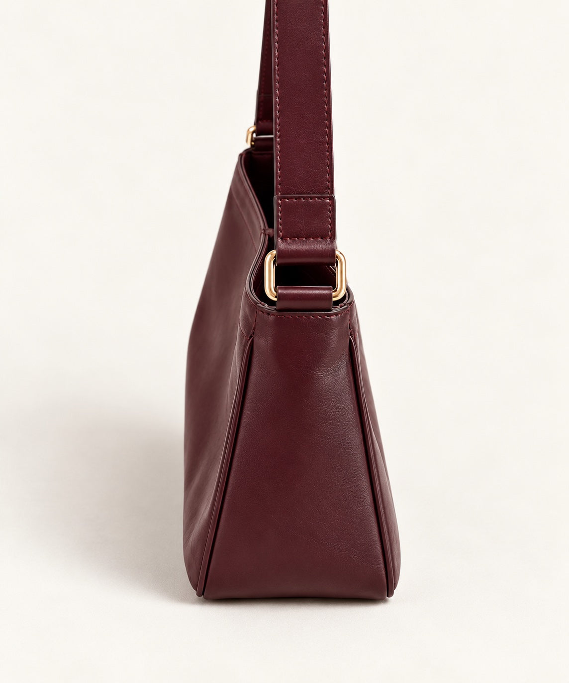 A side view of a dark burgundy leather shoulder bag with gold-tone hardware, set against a clean off-white background.